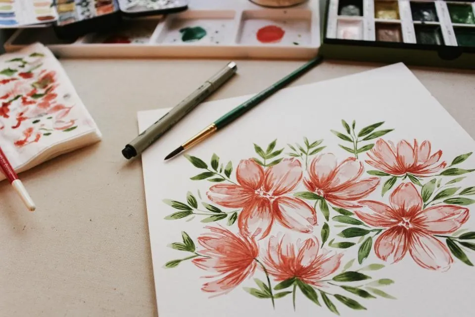Table de l'atelier : une aquarelle de fleurs en cours, des pinceaux, une boîte de couleurs