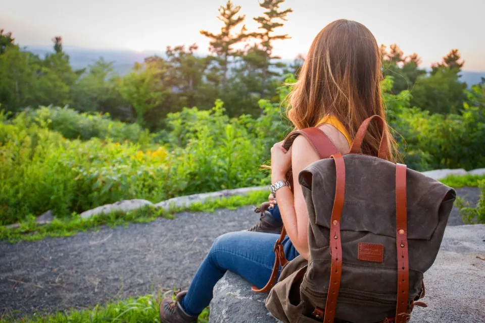 Une randonneuse assise sur un rocher, le sac en toile cirée olive sur le dos, face à la forêt
